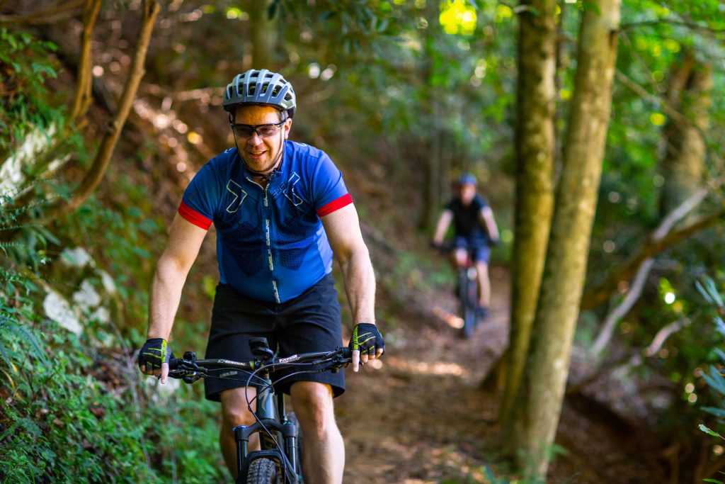 a man on a mountain bike is smiling as he rides down the trail. he is wearing a biking clothes. Behind him is another out-of-focus mountain biker.
