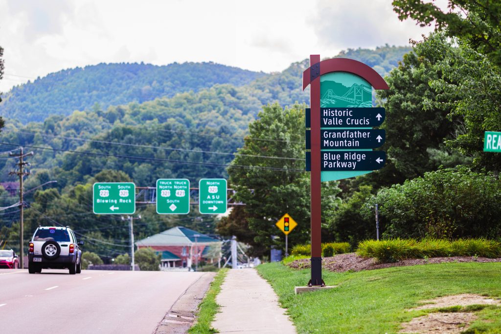 A wayfinding sign is shown by the side of the road in Watauga County. It is a large green sign with blue directional features.