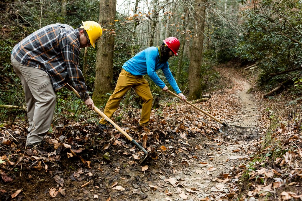 two men wearing hard hats and work clothes work with hoes to build a new trail