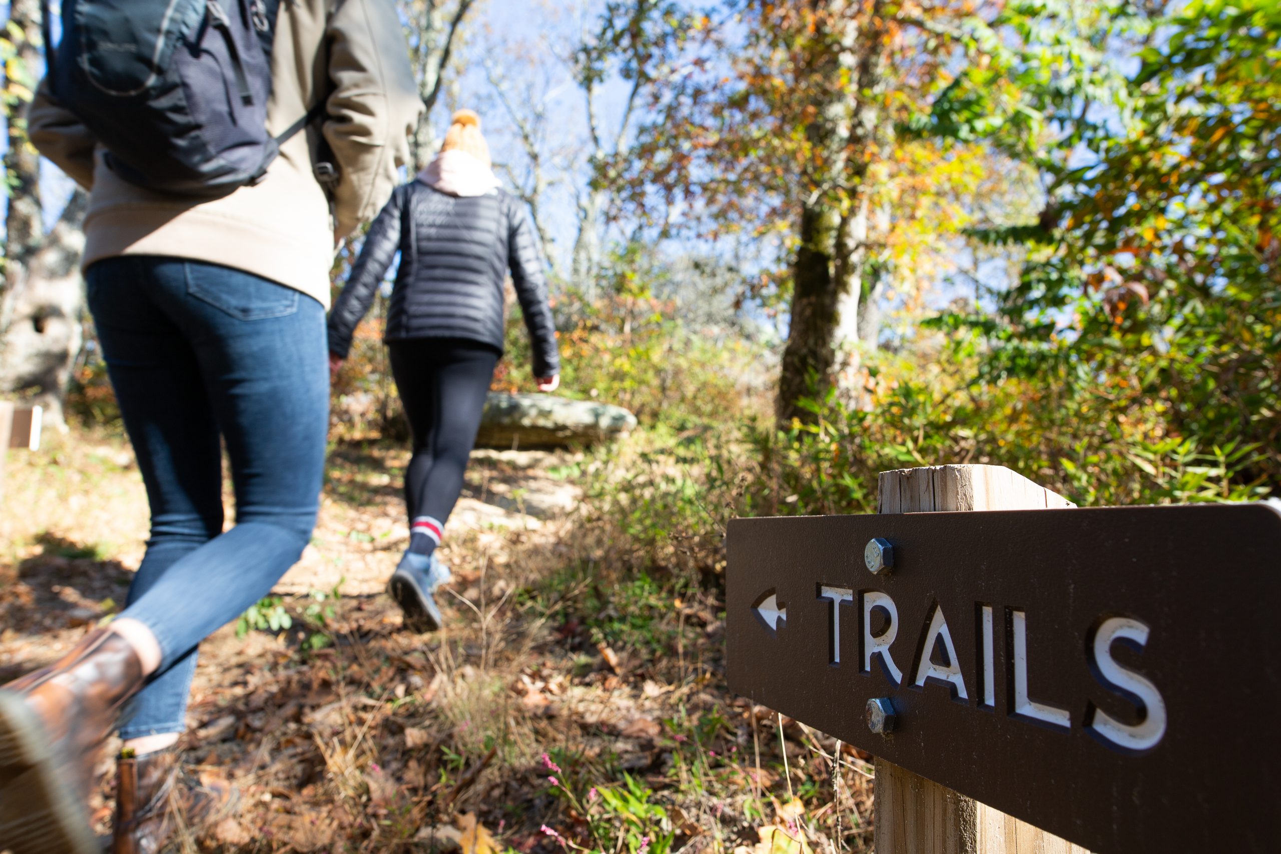 Two women hiking in front of a sign with the word 'trail' and an arrow