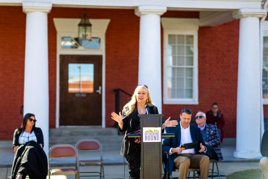A local leader speaks to the crowd at the ribbon cutting ceremony. She has long blonde hair and is wearing a black suit. She is in front of a red brick building with white pillars.