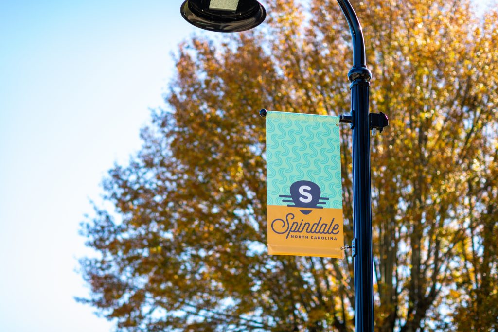 A light post featuring a flag with the Spindale brand. A tree with yellow fall leaves is directly behind the light post.